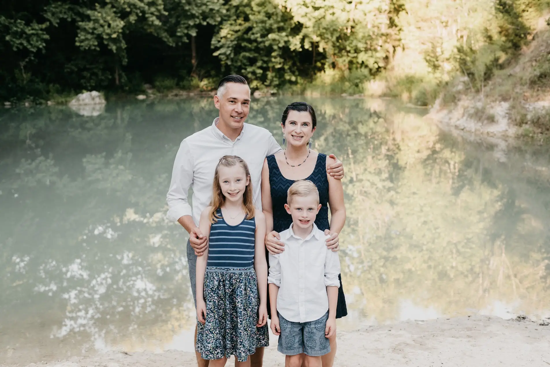 Family of four posing together beside a calm green river surrounded by trees